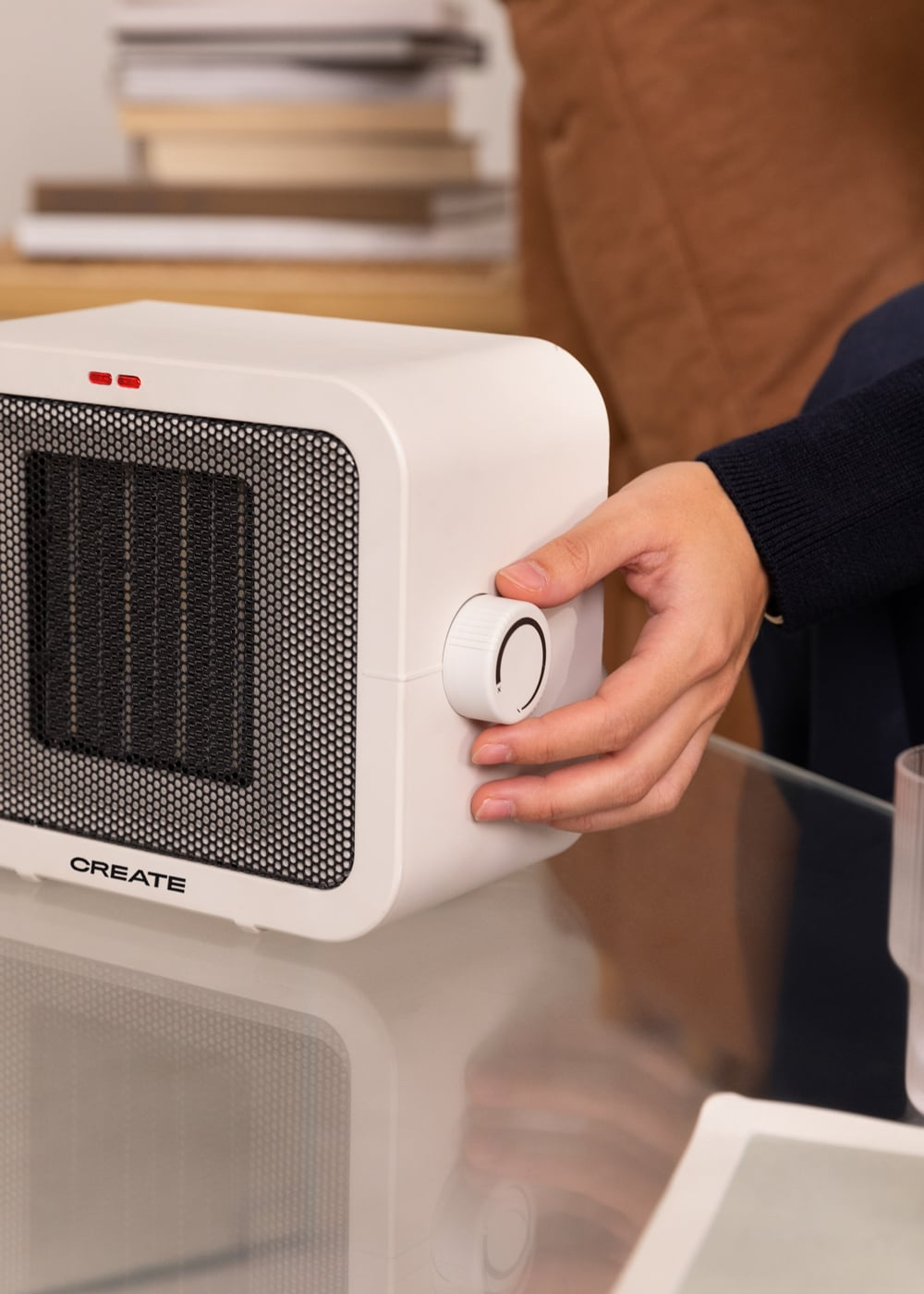 Una mano gira el dial de un calefactor portátil blanco sobre una mesa de cristal. Dos luces rojas están encendidas.