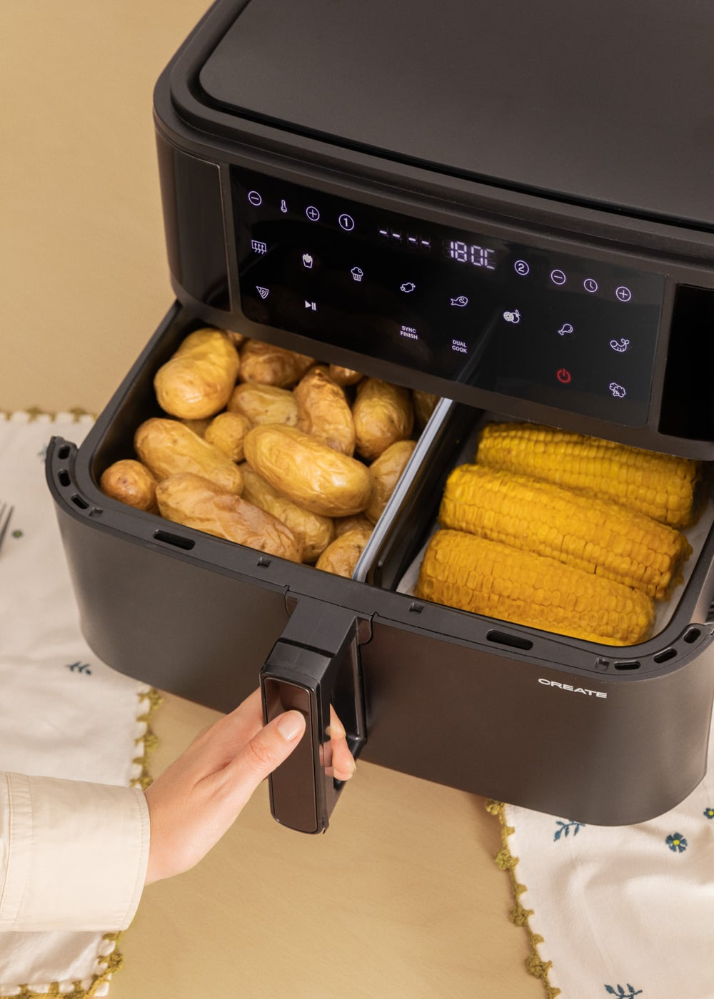 Une main retire le tiroir d'une friteuse à air noire contenant des pommes de terre et des épis de maïs.