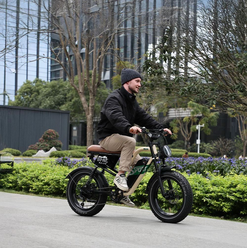 Un hombre monta una bicicleta eléctrica por una acera junto a arbustos verdes y flores moradas.