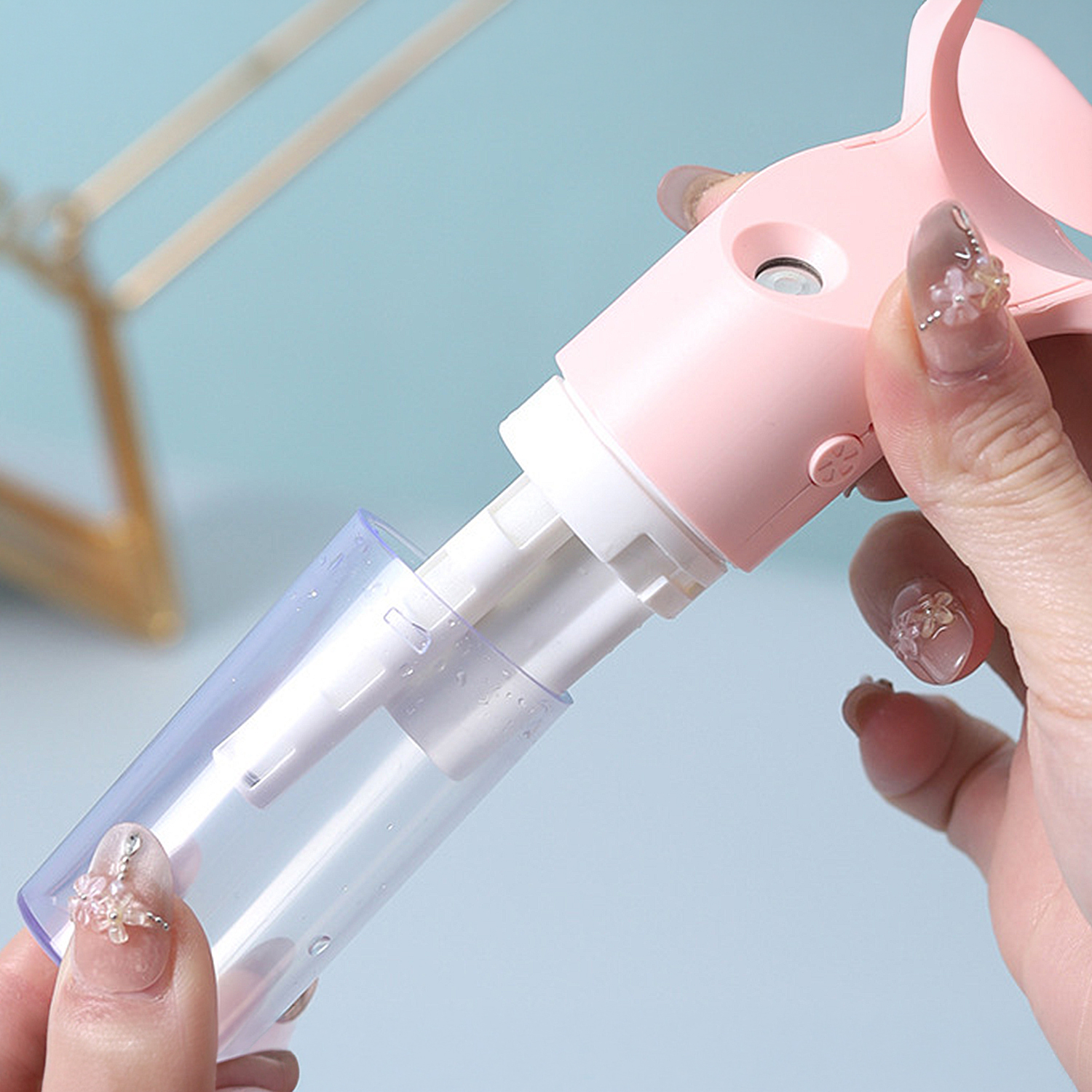 Una mano sostiene un pulverizador rosa con recipiente transparente y partes internas blancas.