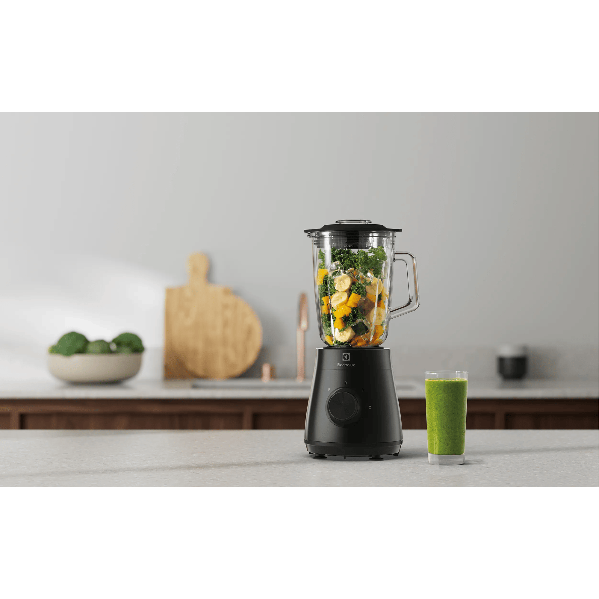 Una licuadora negra con frutas y verduras, al lado un vaso de batido verde.