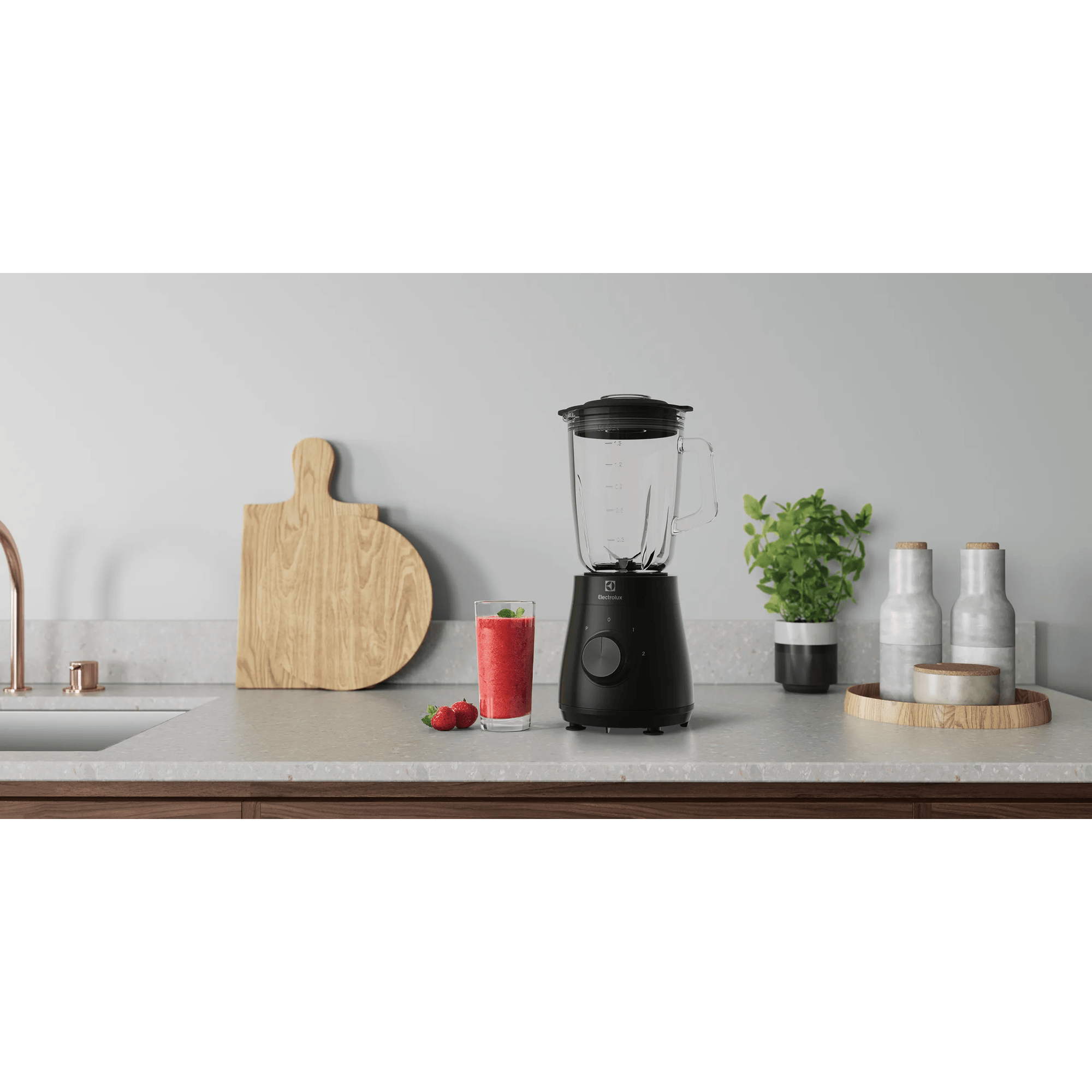 Batidora negra con vaso de batido de fresa, tabla de cortar, planta y tarros.