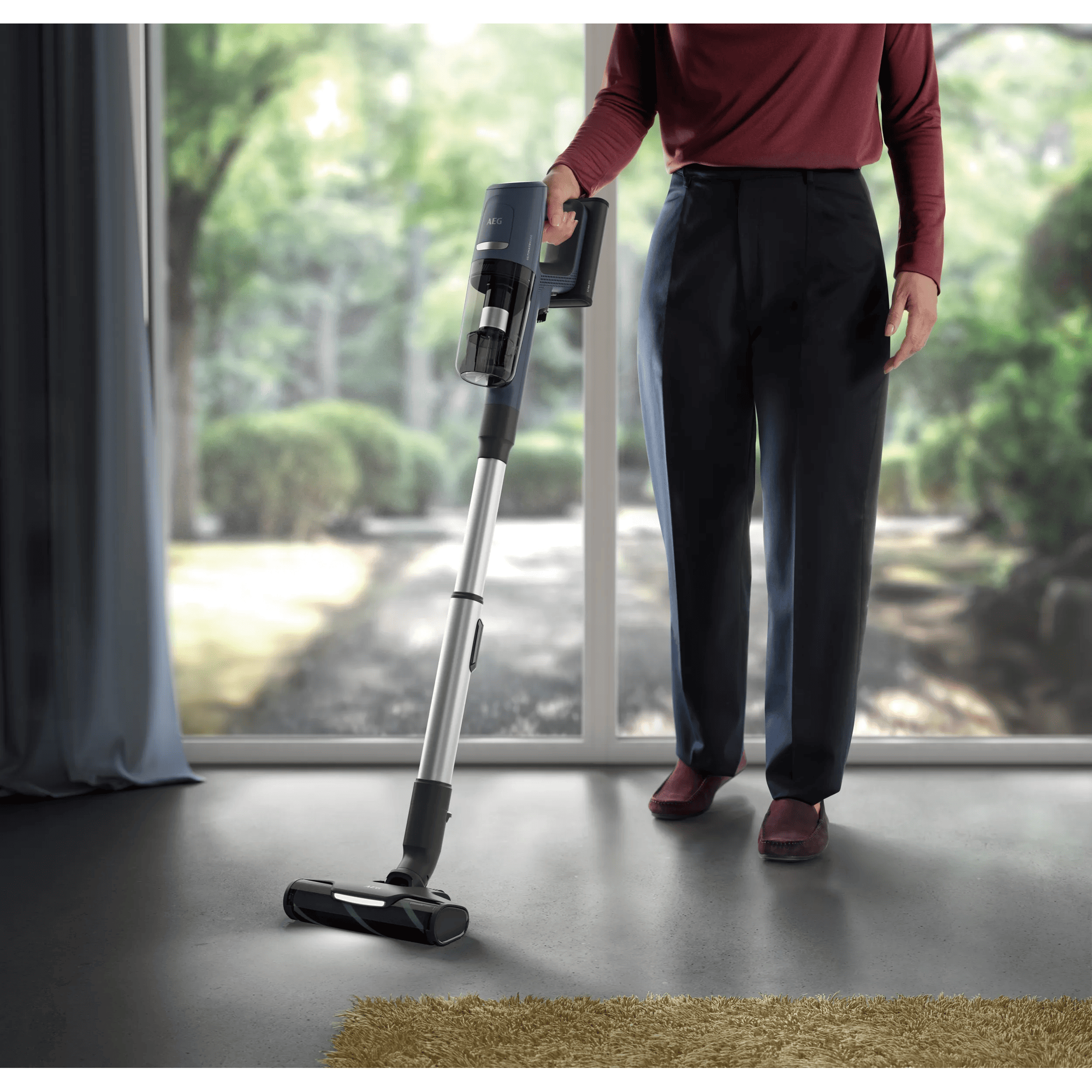 Una persona sostiene una aspiradora inalámbrica azul frente a una gran ventana con vistas a un jardín.