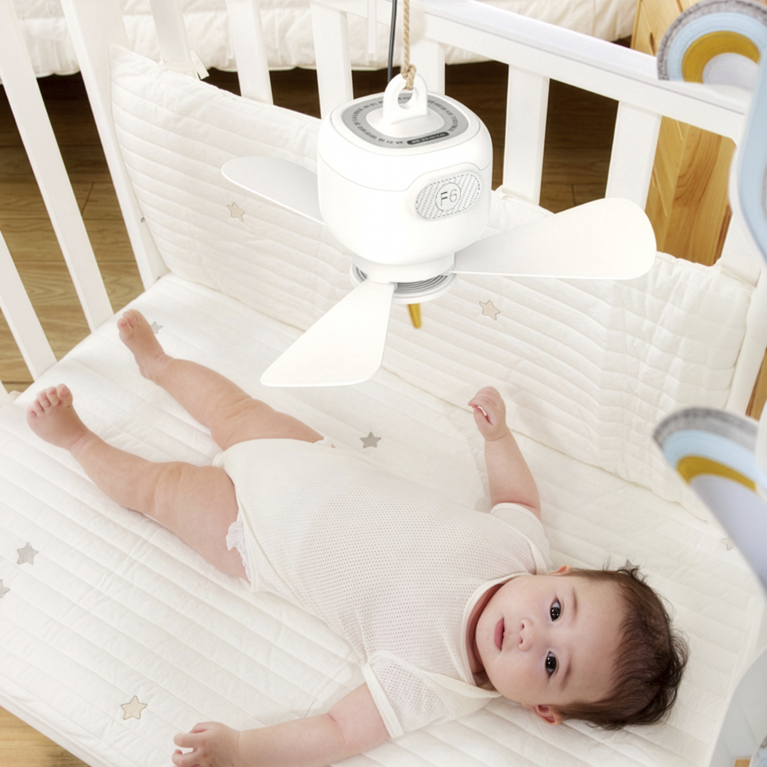 Bebé en mameluco blanco yace en una alfombra de cuna blanca con un ventilador blanco colgando encima.