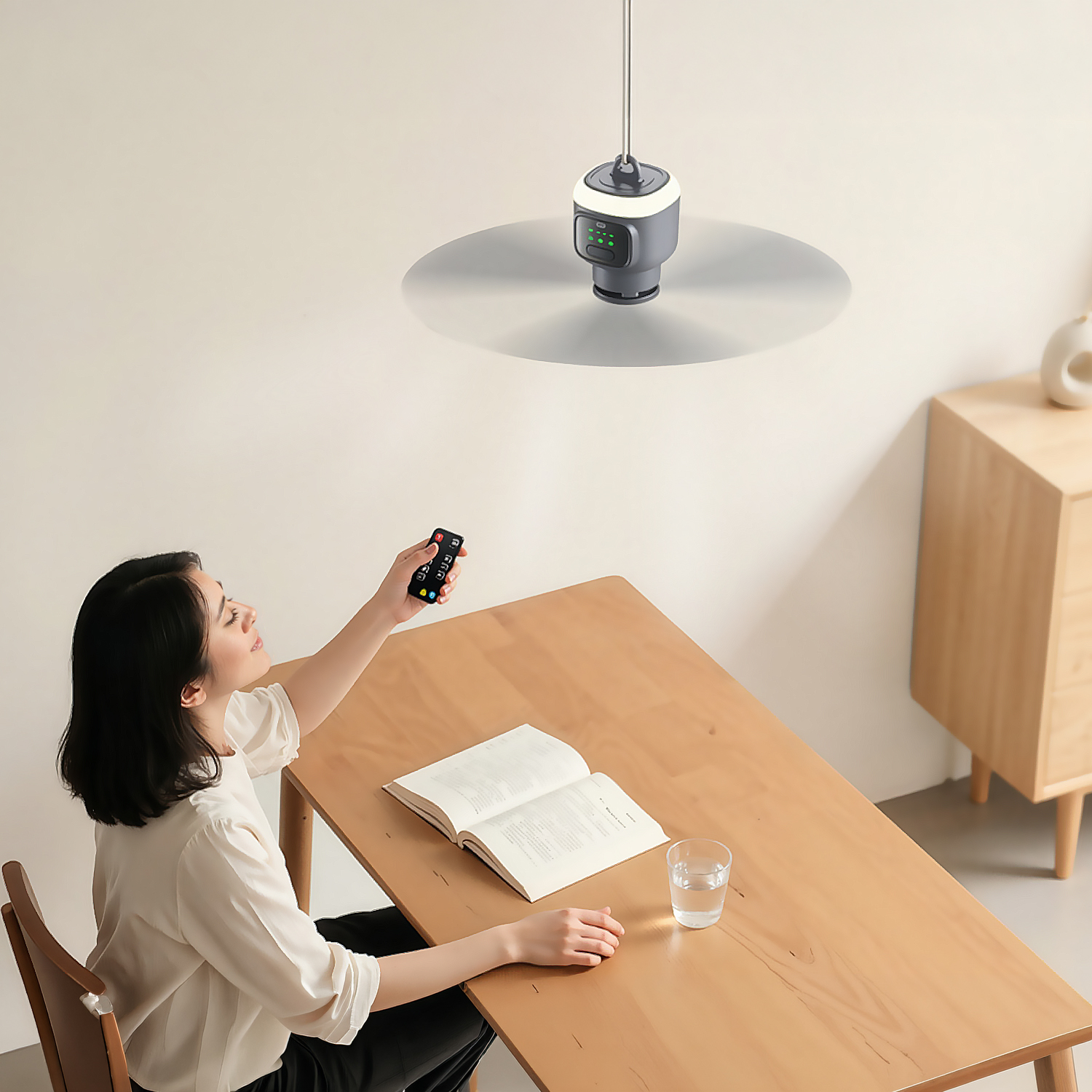 Mujer en mesa con control remoto, libro y vaso. Luz de ventilador de techo arriba.
