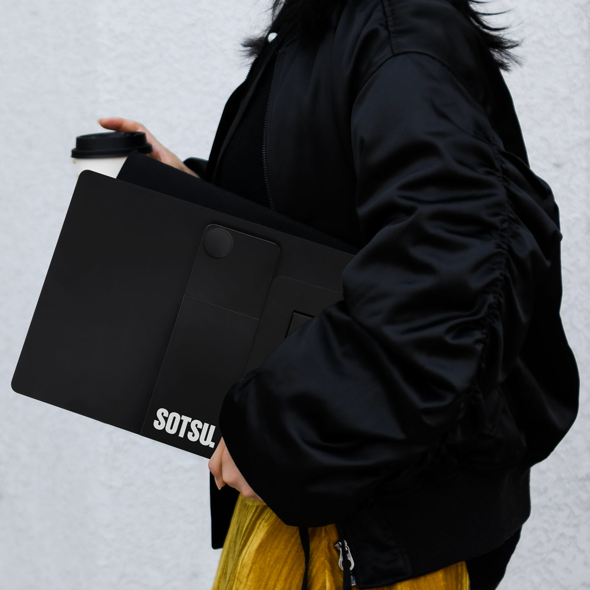 Osoba w czarnej kurtce trzymająca czarny laptop i kubek kawy. Żółta spódnica.