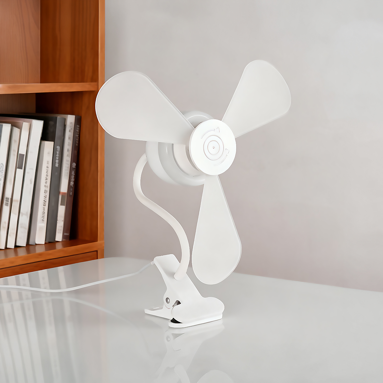 Un ventilador blanco con pinza y tres aspas está sobre un escritorio blanco junto a una estantería de madera.