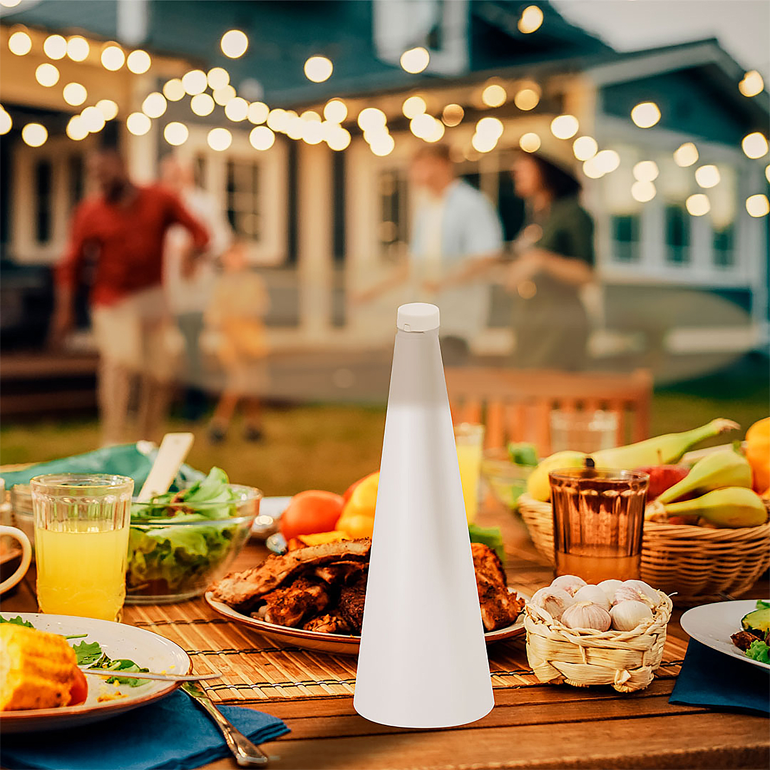 Mesa al aire libre con comida, bebidas y luces; gente borrosa al fondo.
