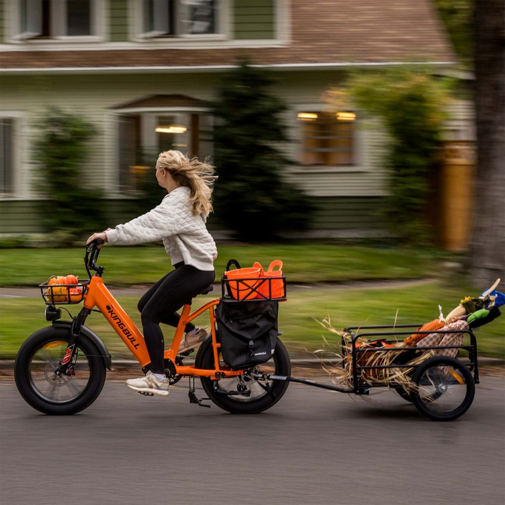 Mujer monta bicicleta naranja con remolque lleno de calabazas y espantapájaros.