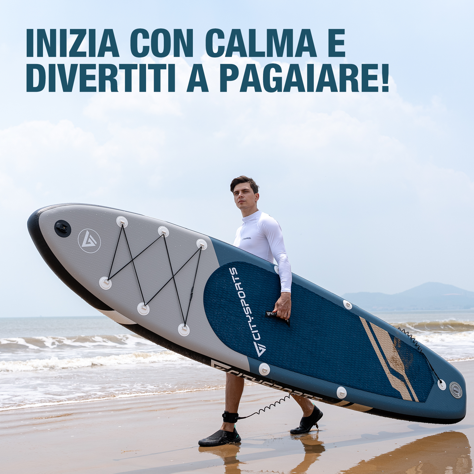 Un uomo in camicia bianca porta una tavola da paddle blu e grigia sulla spiaggia.