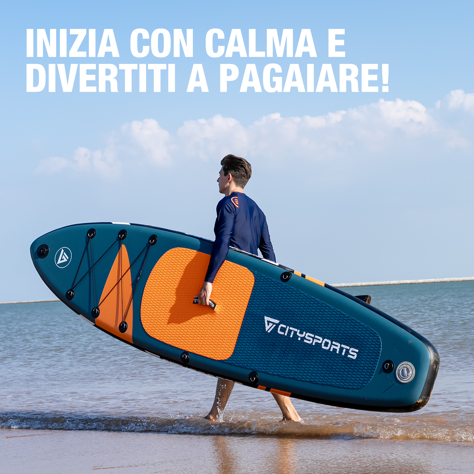 Un uomo con maglia blu a maniche lunghe porta una tavola da SUP blu e arancione sulla spiaggia.
