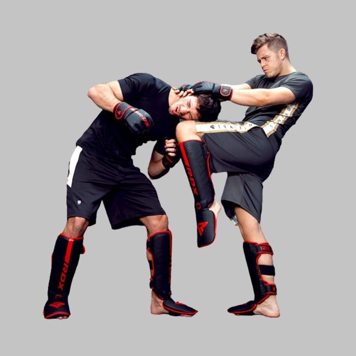 Two men sparring in MMA gear. One kicks the other in the head while holding his neck.