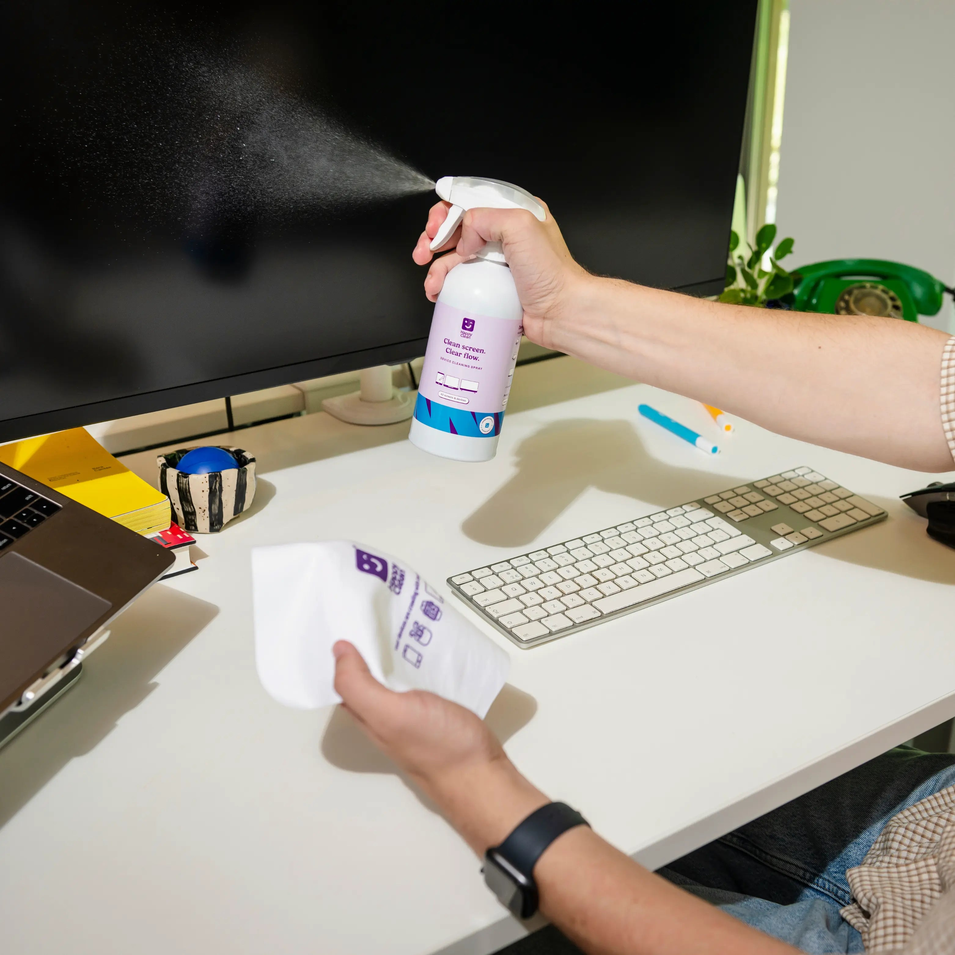 Hand spuit schermreiniger op een monitor op een bureau met toetsenbord en laptop.