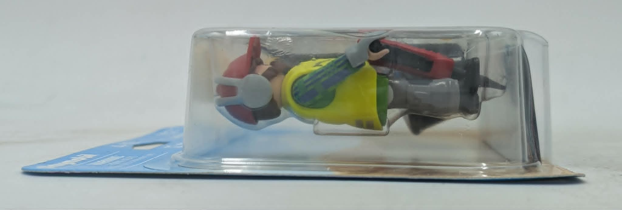 Eine Spielzeugfigur in einer Plastikverpackung. Sie trägt einen gelben Körper und rote und blaue Akzente.