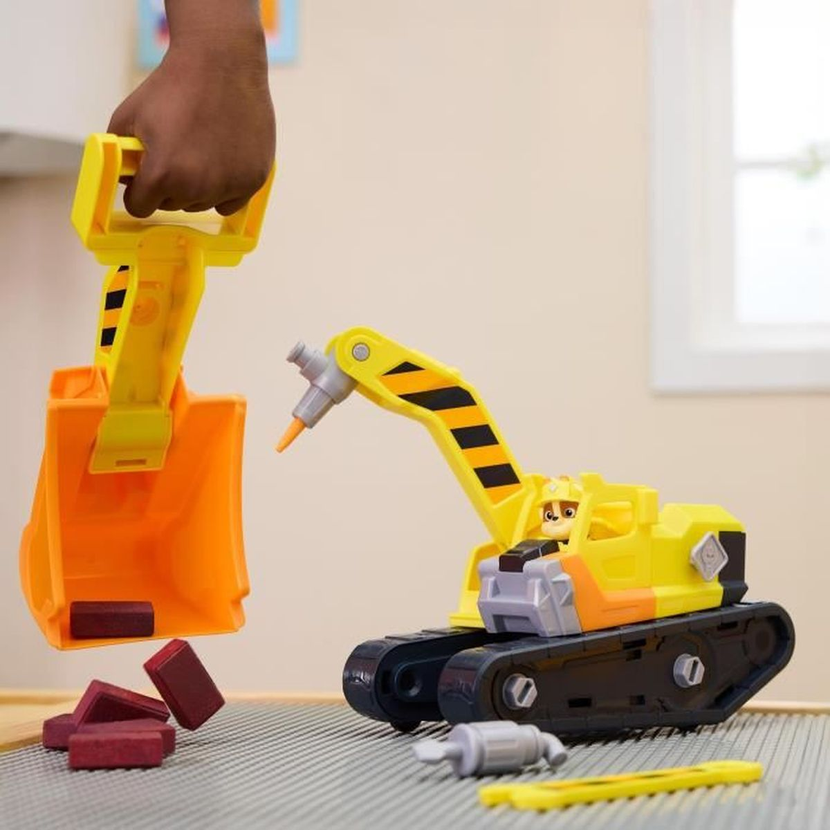 Una mano infantil sostiene una pala de juguete naranja sobre ladrillos de juguete y una excavadora de juguete amarilla.