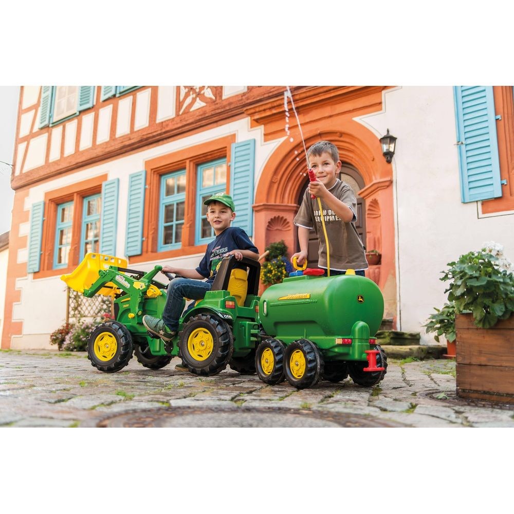 Zwei Jungen auf einem grünen Spielzeugtraktor mit Anhänger vor einem Haus.