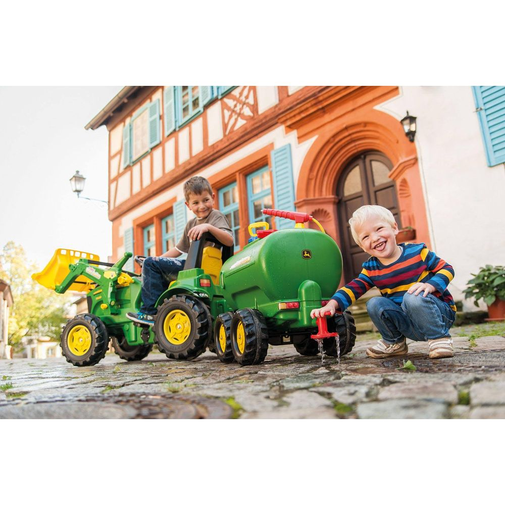 Zwei Jungen spielen mit einem grünen Spielzeugtraktor und einem Wassertank vor einem Haus.
