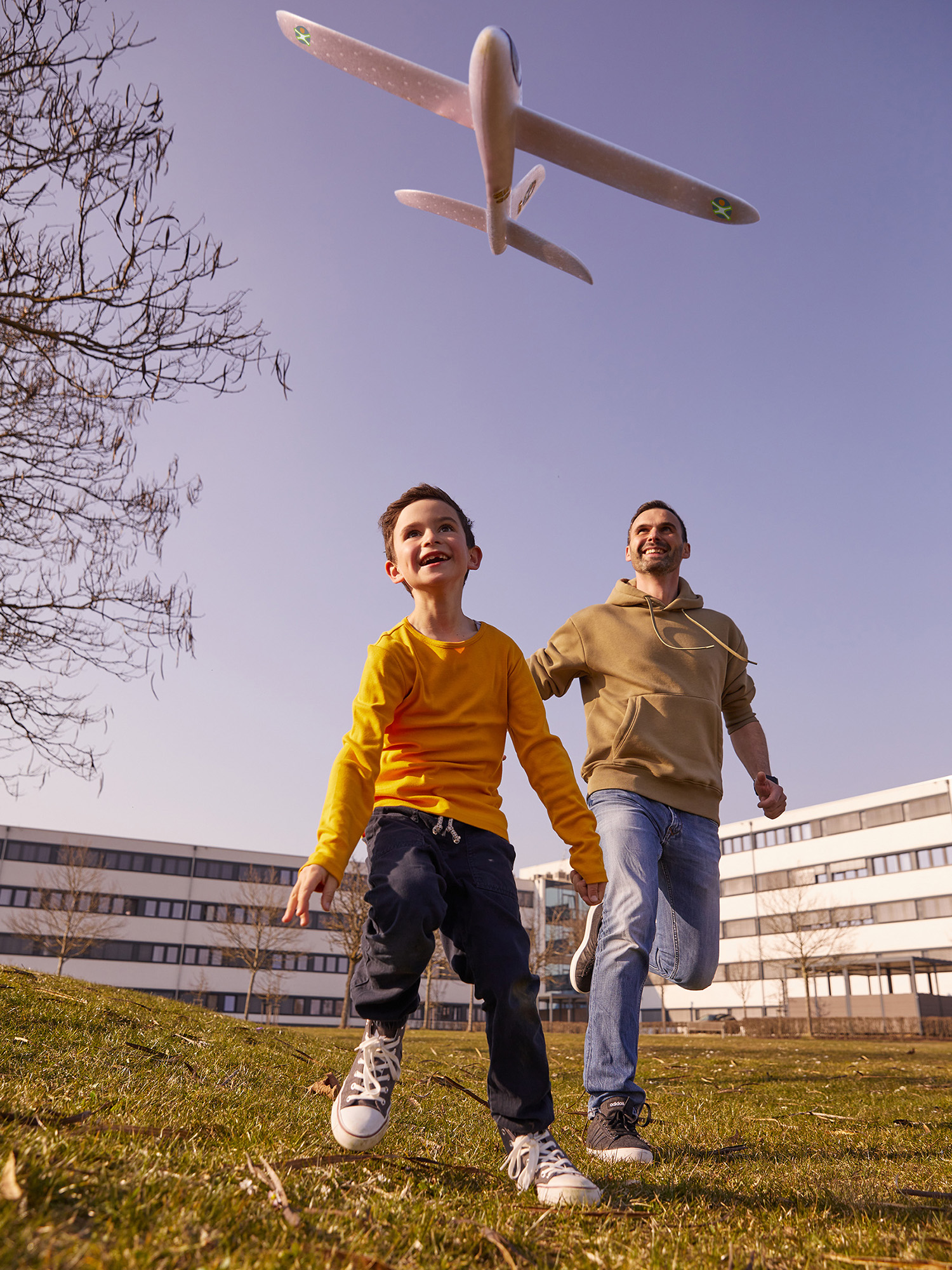 Ein Junge und ein Mann rennen auf einer Wiese unter einem Flugzeug am Himmel.