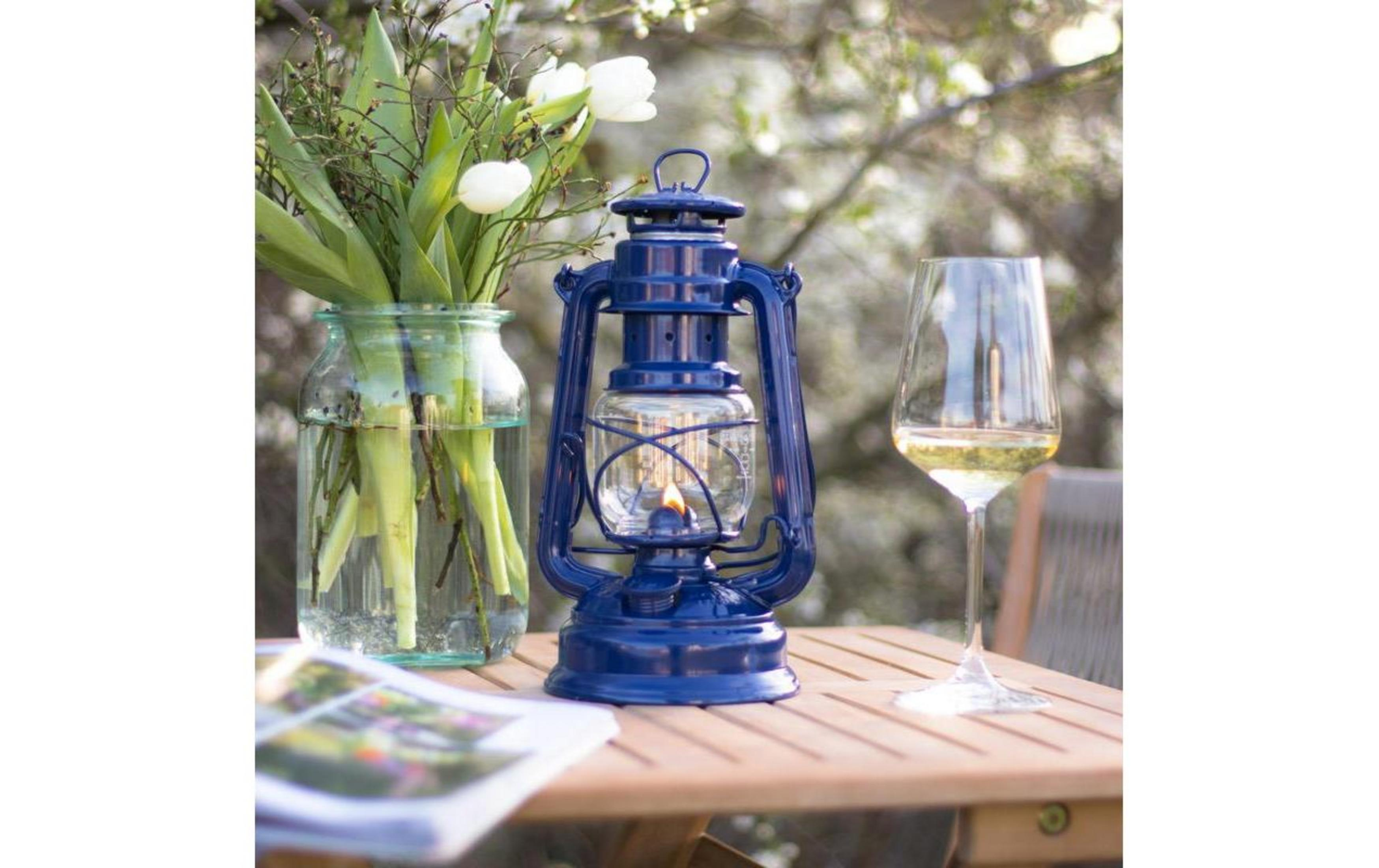 Eine blaue Laterne brennt neben einem Glas Wein und Blumen auf einem Holztisch.