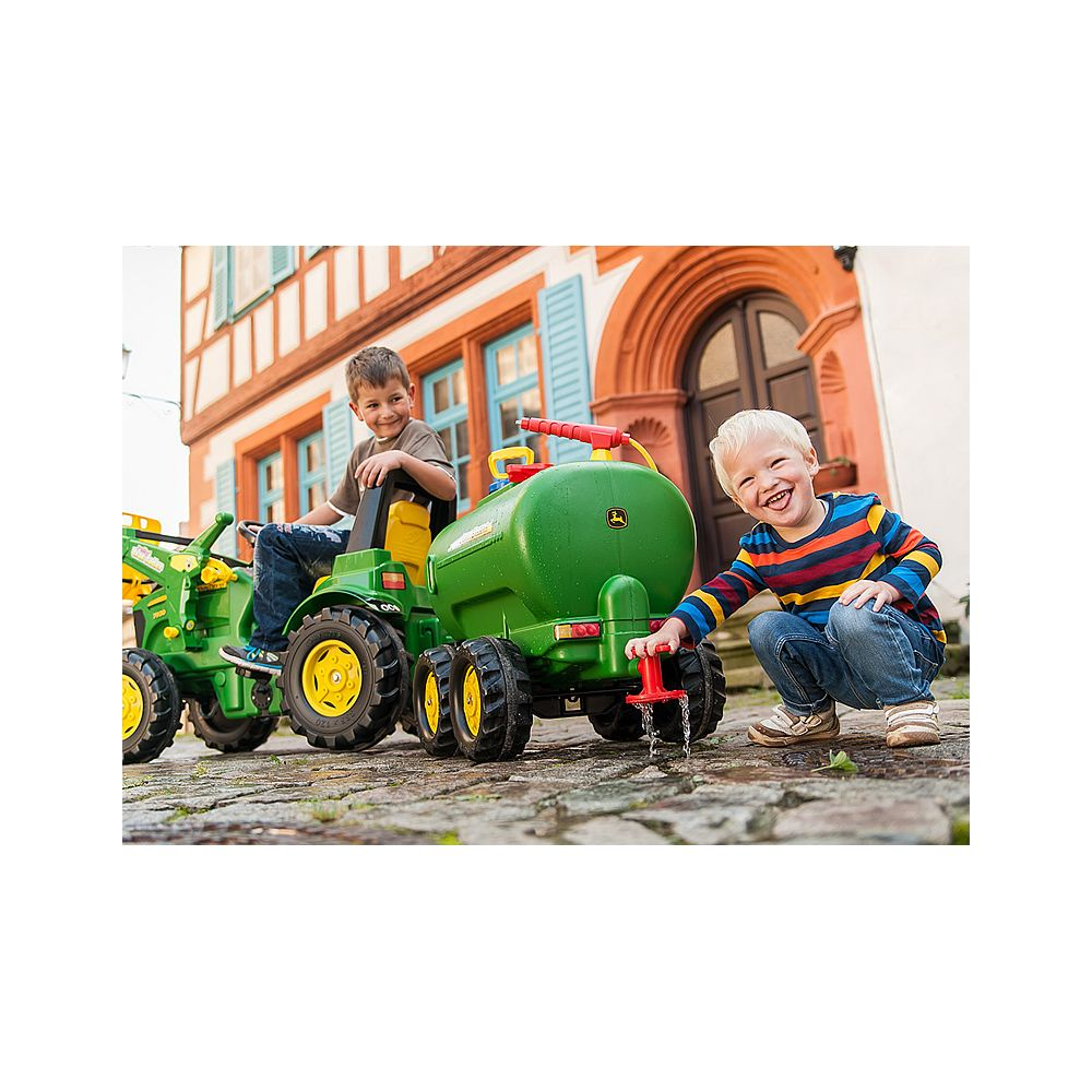 Zwei Jungen spielen mit einem grünen Spielzeug-Traktor und einem Wasserwagen vor einem Haus.