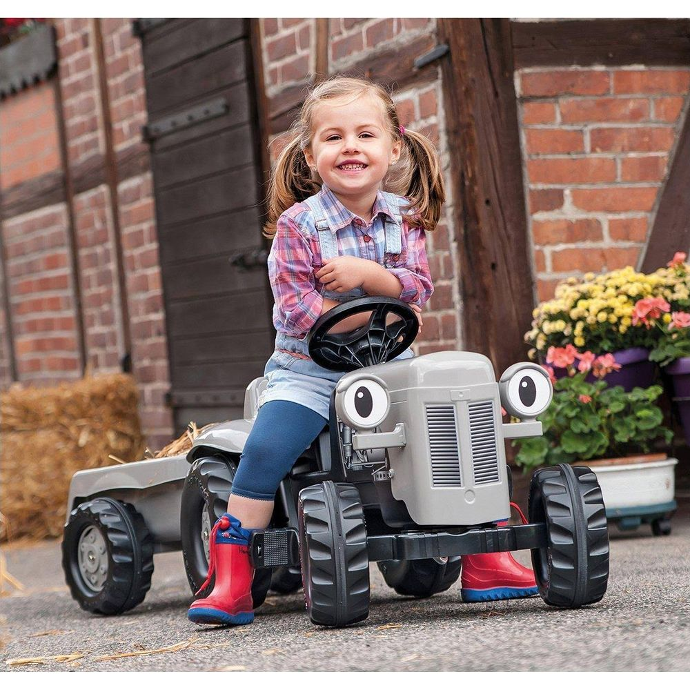 Ein kleines Mädchen sitzt auf einem Spielzeugtraktor mit Anhänger vor einem Bauernhaus.