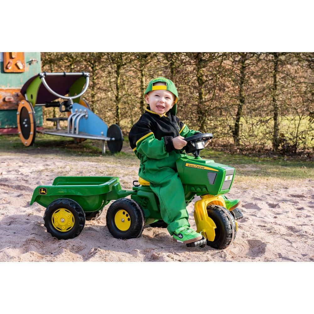 Ein Kleinkind in einem grünen John Deere Anzug fährt einen grünen Spielzeugtraktor mit Anhänger.
