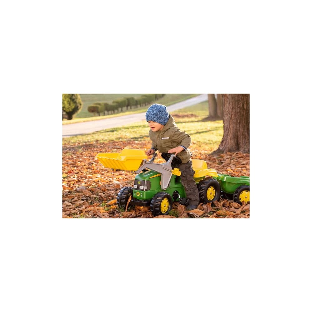 Ein Junge in einer blauen Mütze fährt einen grünen Spielzeugtraktor mit Anhänger im Herbstlaub.