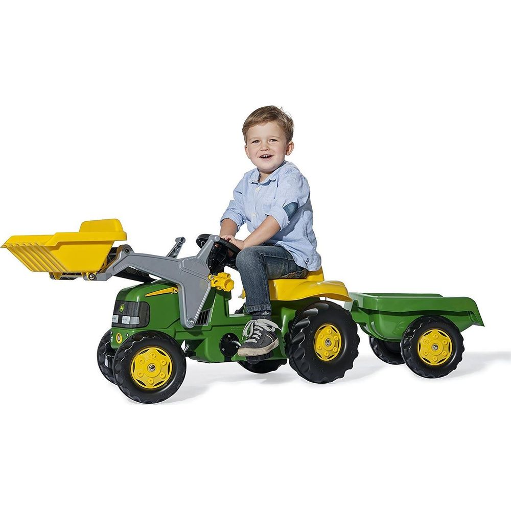 Ein Junge sitzt auf einem grünen Spielzeugtraktor mit Lader und Anhänger.