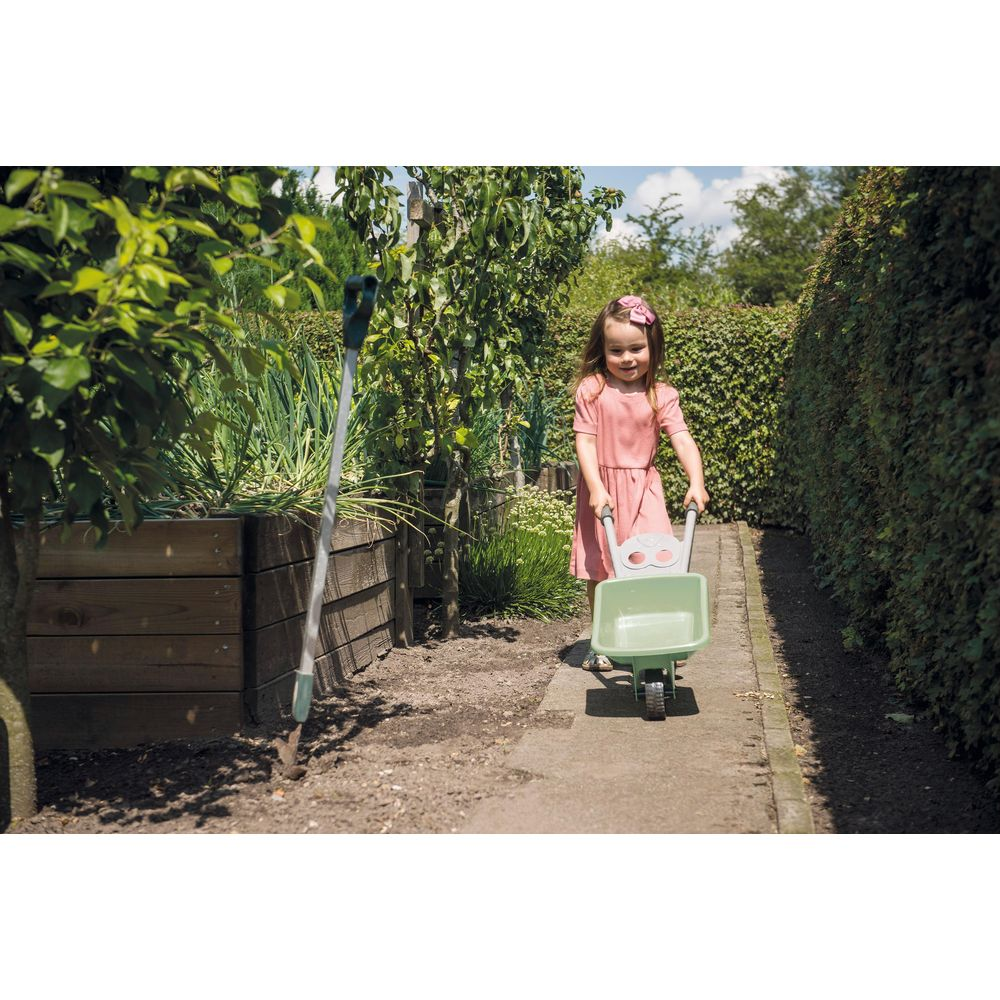 Ein junges Mädchen im rosa Kleid schiebt eine grüne Spielzeugschubkarre auf einem Gartenweg.