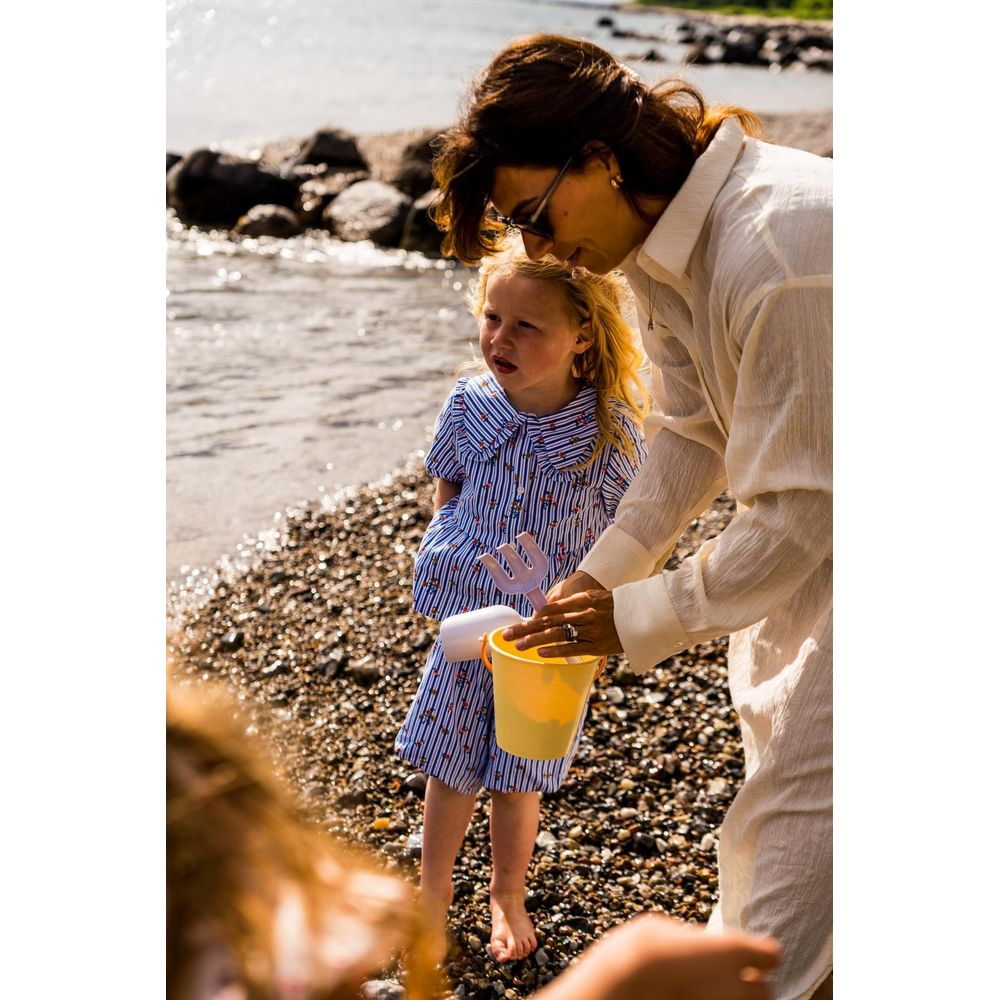 Mutter hilft Kind mit gelbem Eimer und Spielzeug am Kiesstrand.