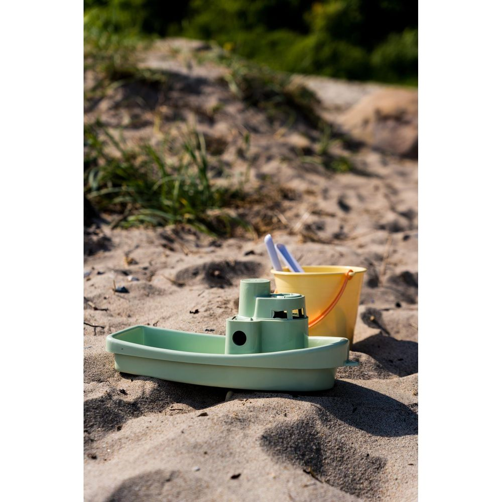 Ein grünes Spielzeugboot und ein gelber Eimer stehen auf Sand. Grünes Gras im Hintergrund.