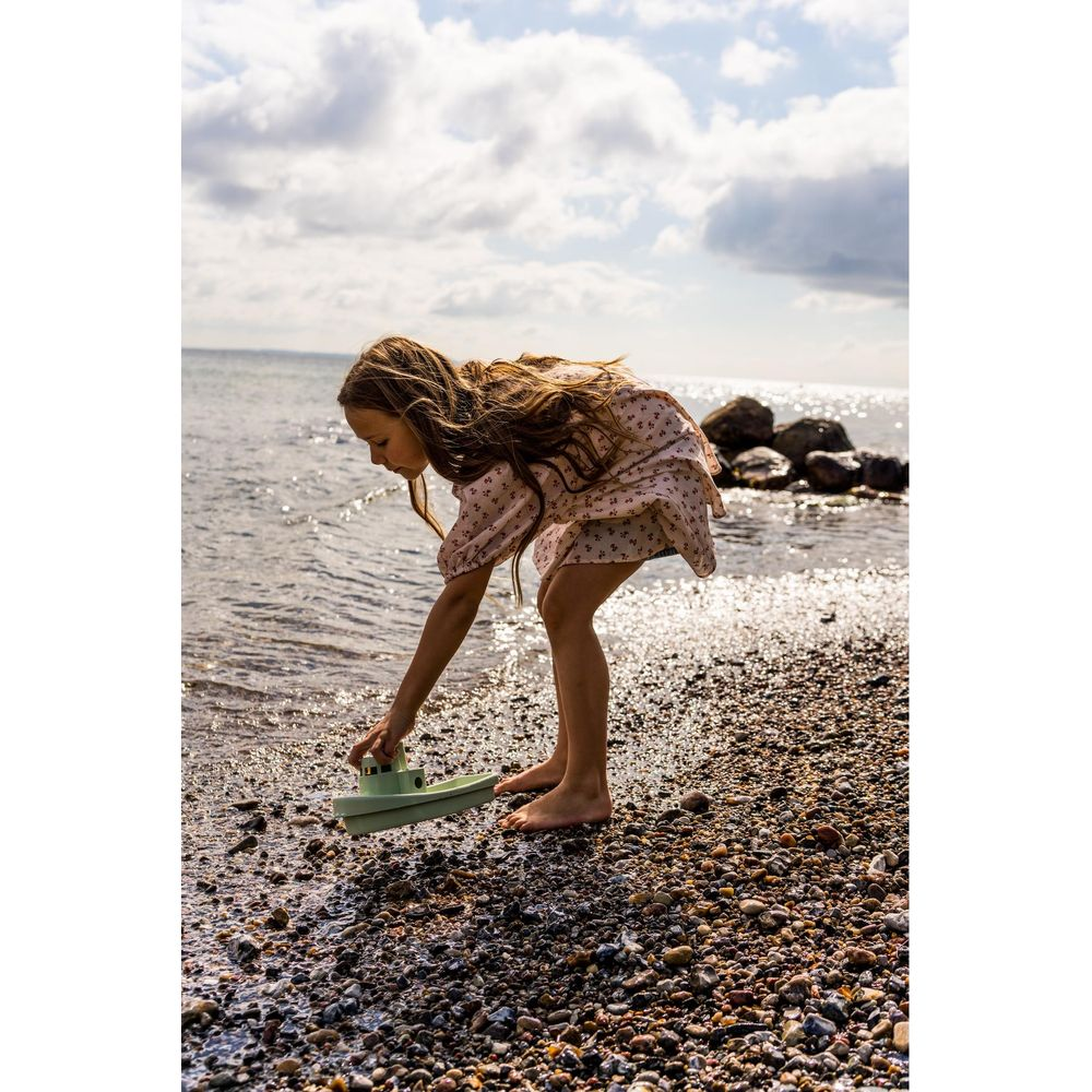 Ein Mädchen spielt mit einem grünen Spielzeugboot am steinigen Strand.