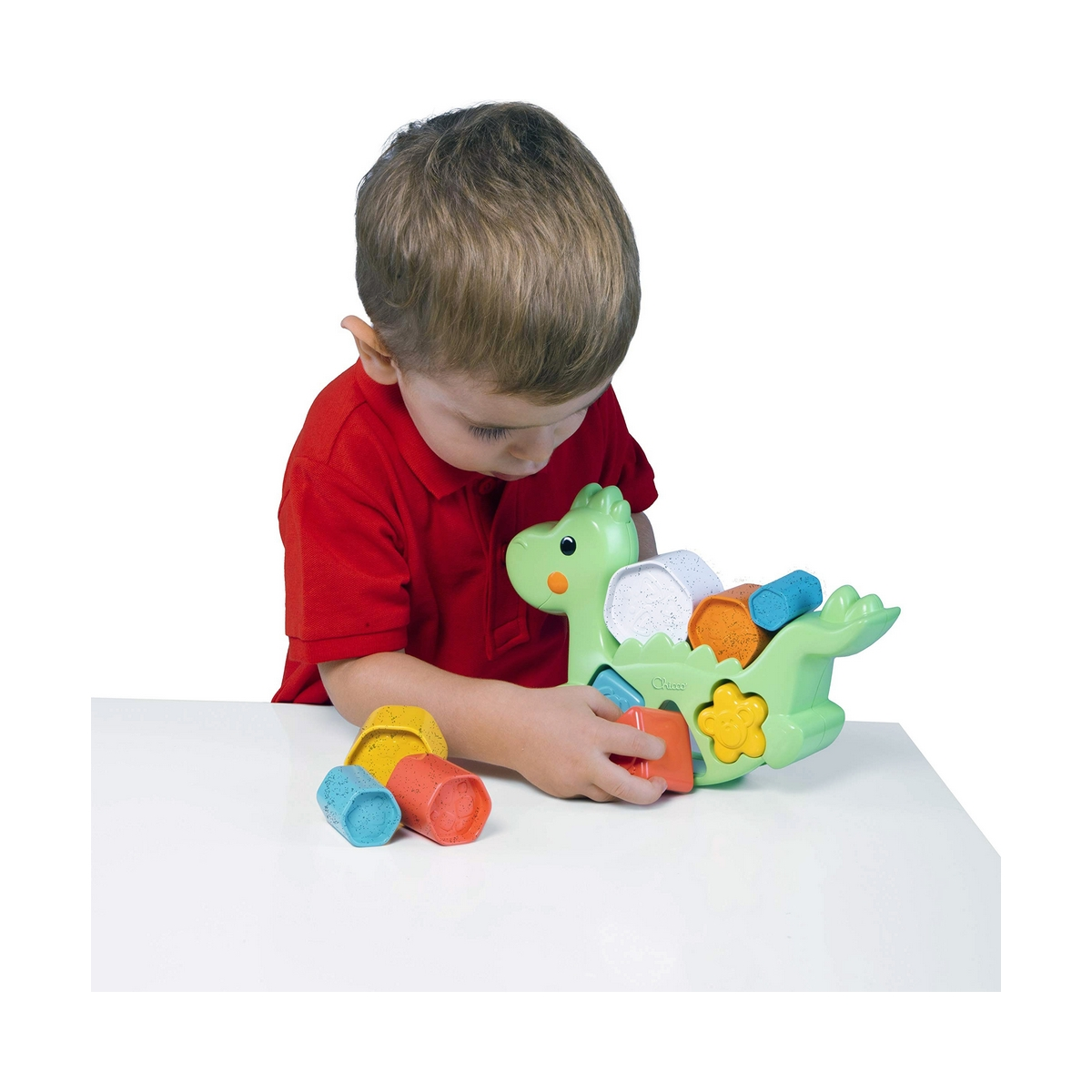 Ein kleiner Junge in einem roten Hemd spielt mit einem grünen Dinosaurierspielzeug auf einem weißen Tisch.