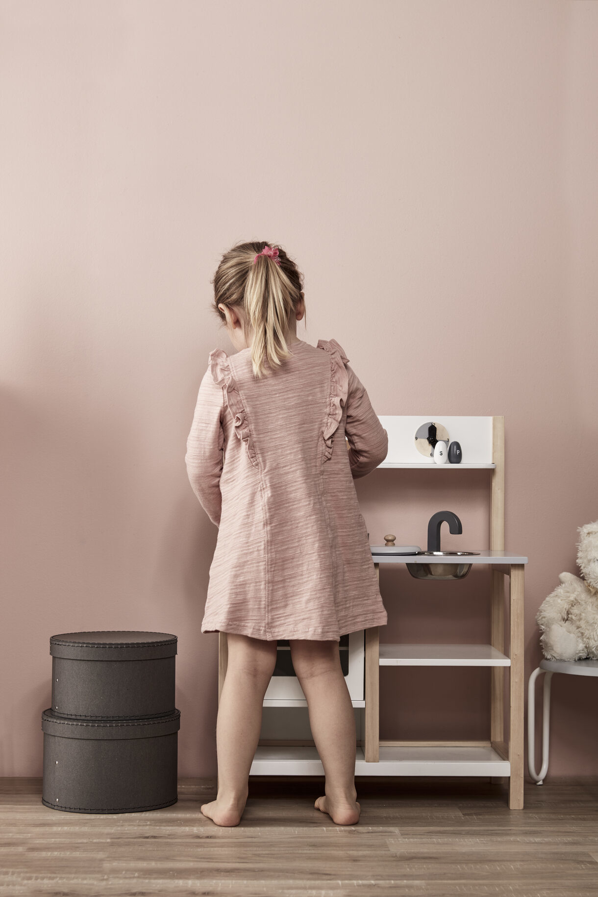 Ein kleines Mädchen in rosa spielt mit einer Spielküche, daneben graue Boxen.