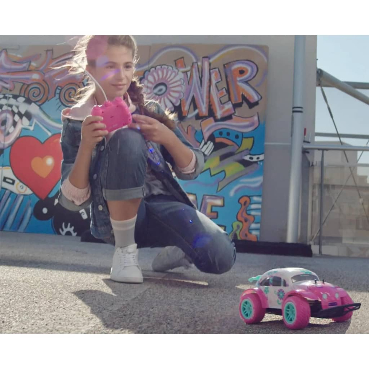 Mädchen mit rosa Kamera kniet neben Spielzeugauto vor Graffiti.