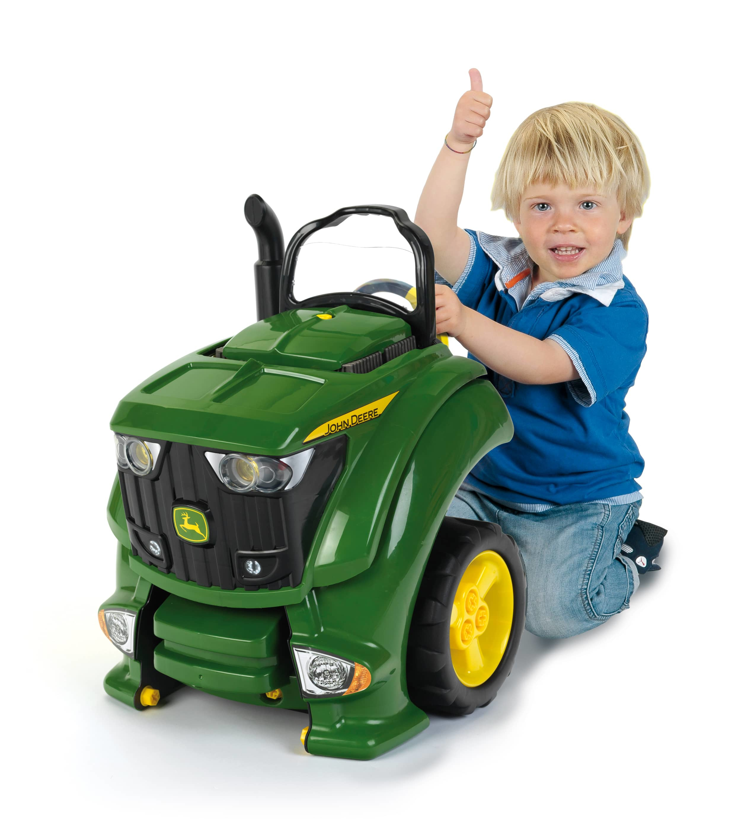 Un niño de pelo rubio da pulgares arriba junto a un tractor de juguete John Deere verde.