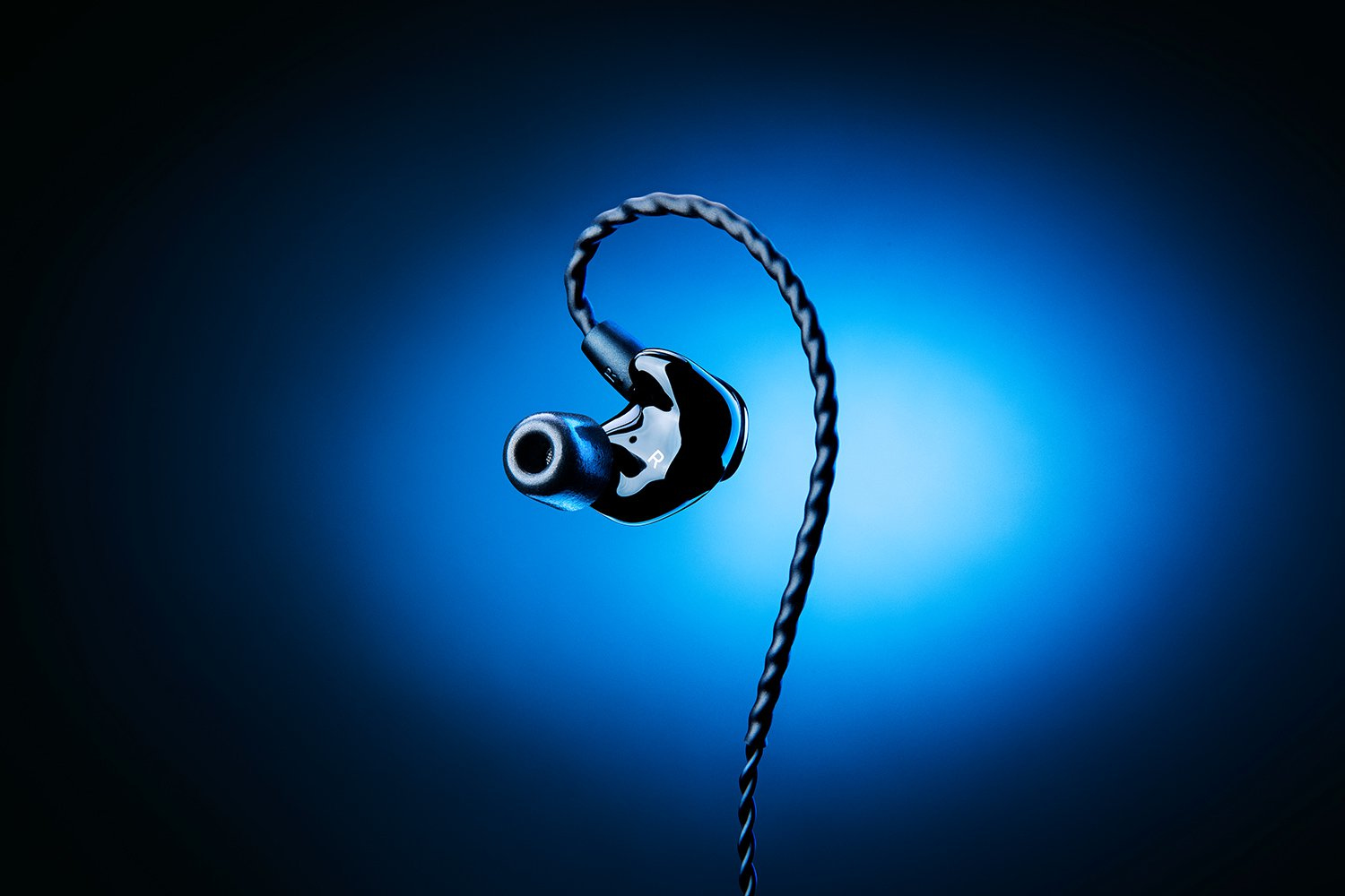 Un auricolare in-ear monitor nero lucido con cavo intrecciato su sfondo blu scuro.