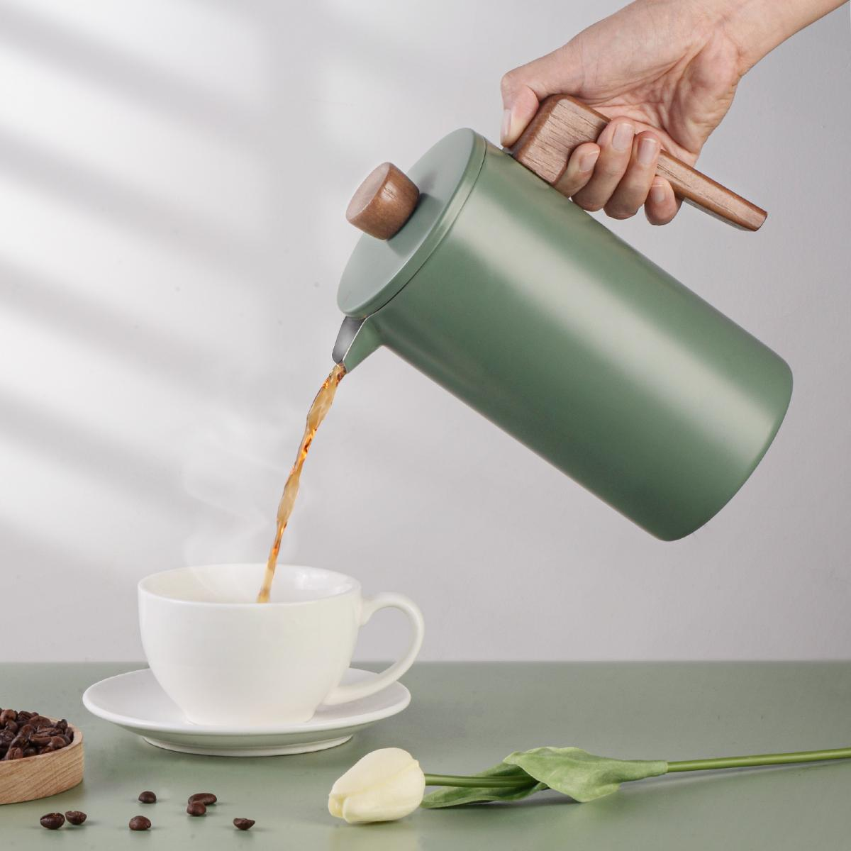 Eine grüne Kanne mit Holzgriff gießt Kaffee in eine weiße Tasse.