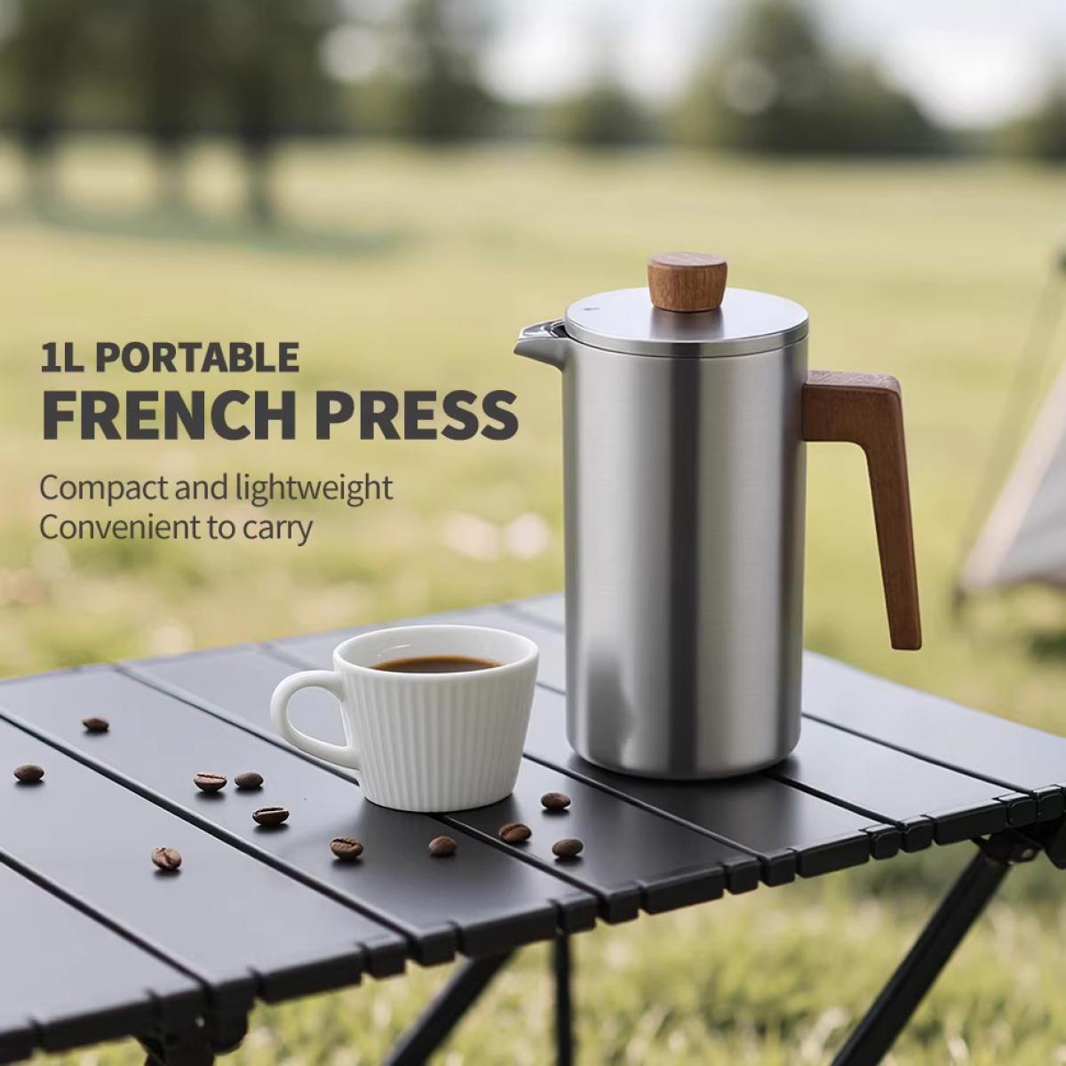 Eine silberne French Press mit Holzgriff und Deckel steht neben einer weißen Tasse Kaffee auf einem Tisch.