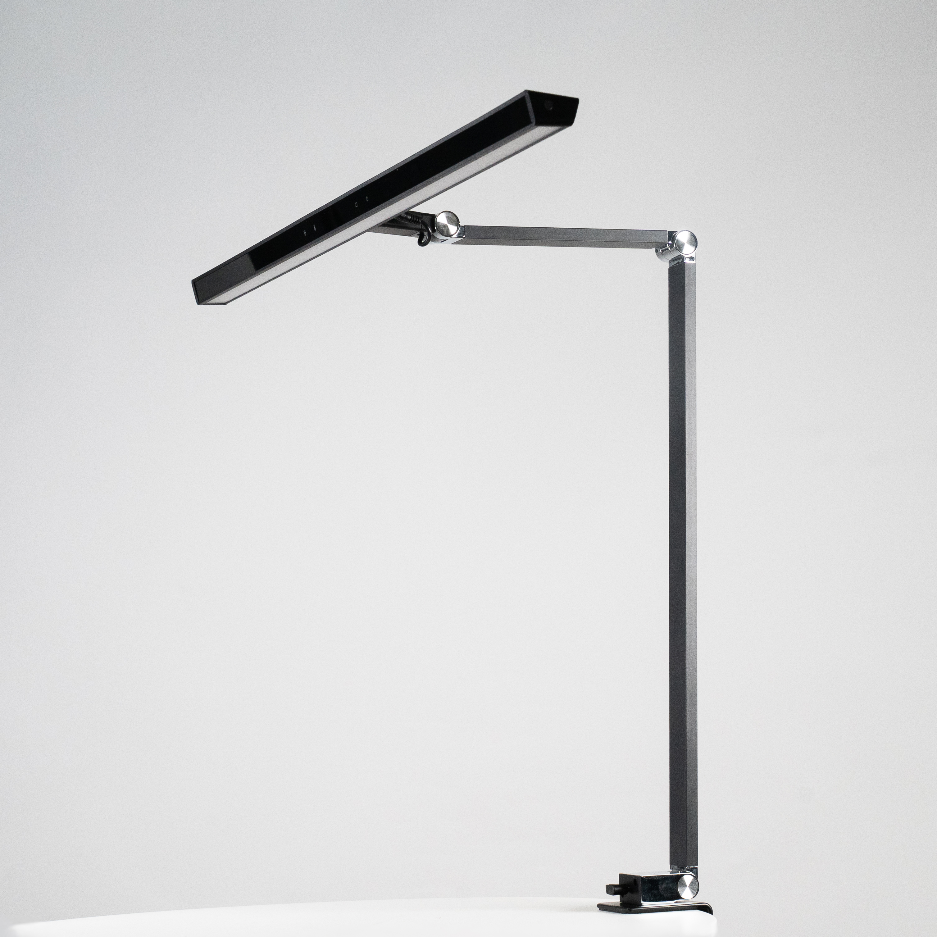 Ein moderner schwarzer Schreibtischarm mit LED-Lichtleiste auf einem weißen Tisch.