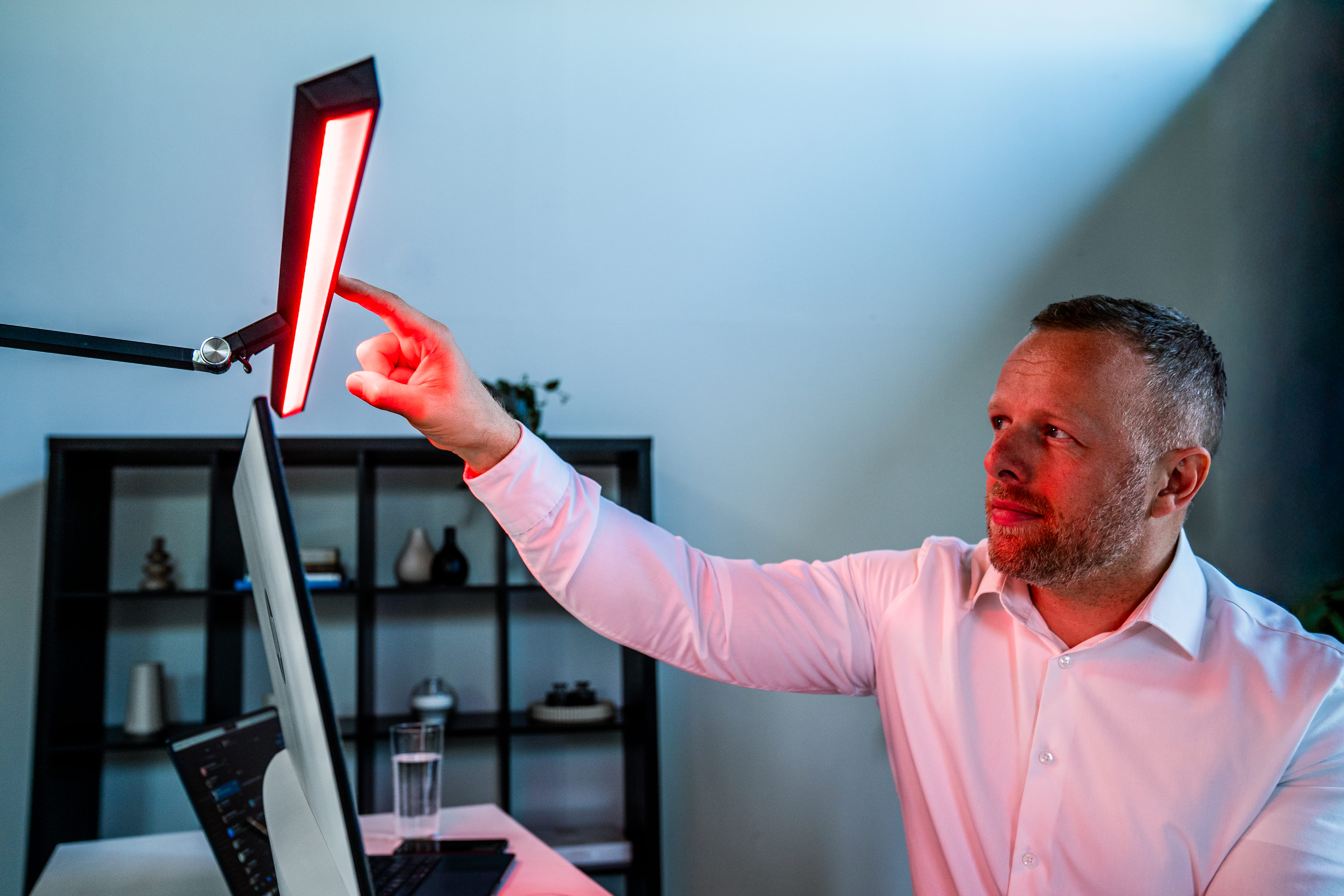 Mann im weißen Hemd berührt rote Lampe über Computertisch. Regal im Hintergrund.