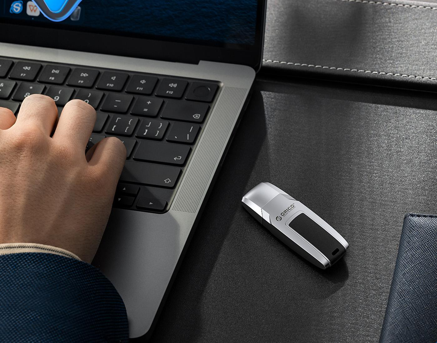 Bir el dizüstü bilgisayarda yazıyor, yanında USB bellek ve defter var.