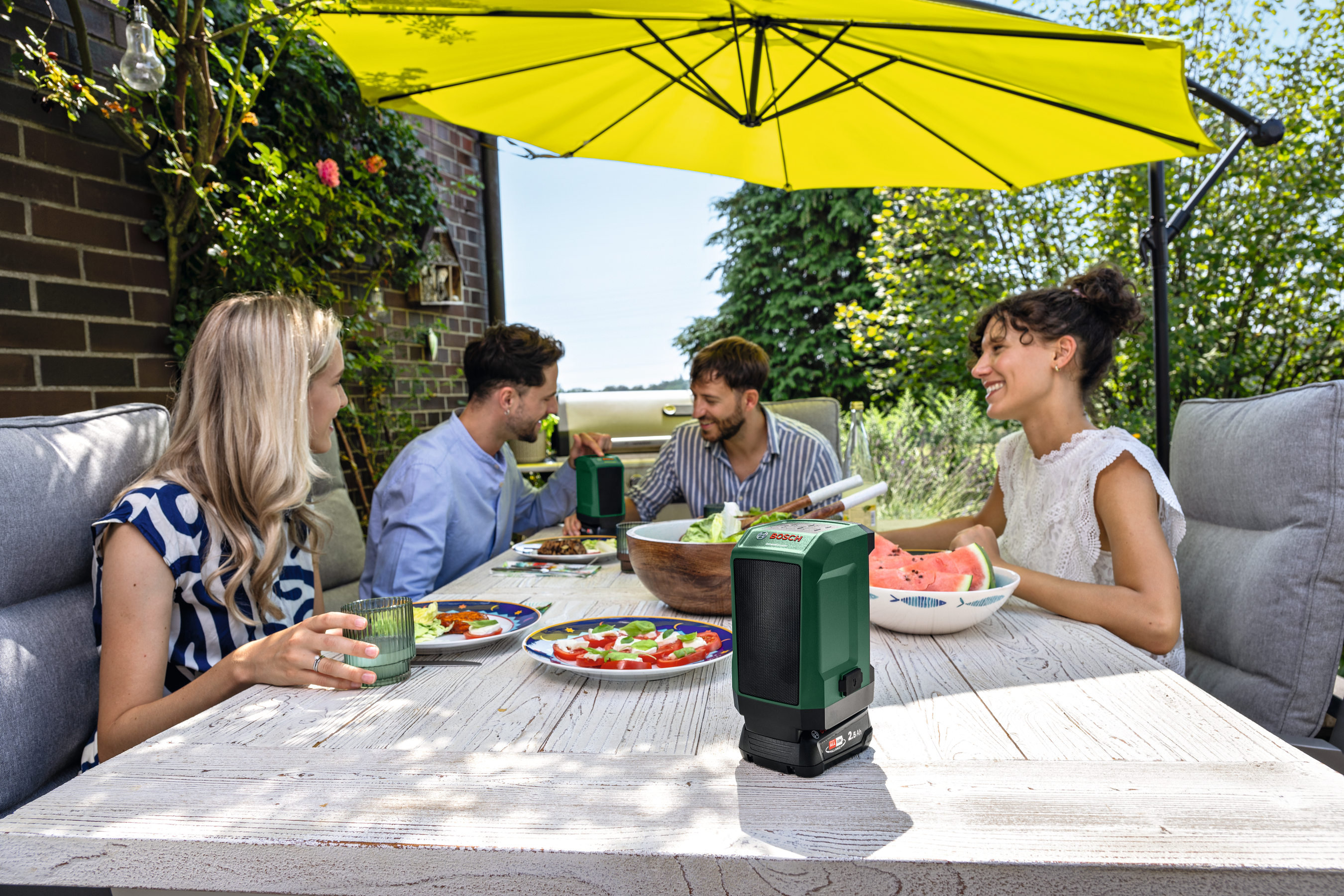 Vier Personen genießen ein Essen unter einem gelben Sonnenschirm im Garten.