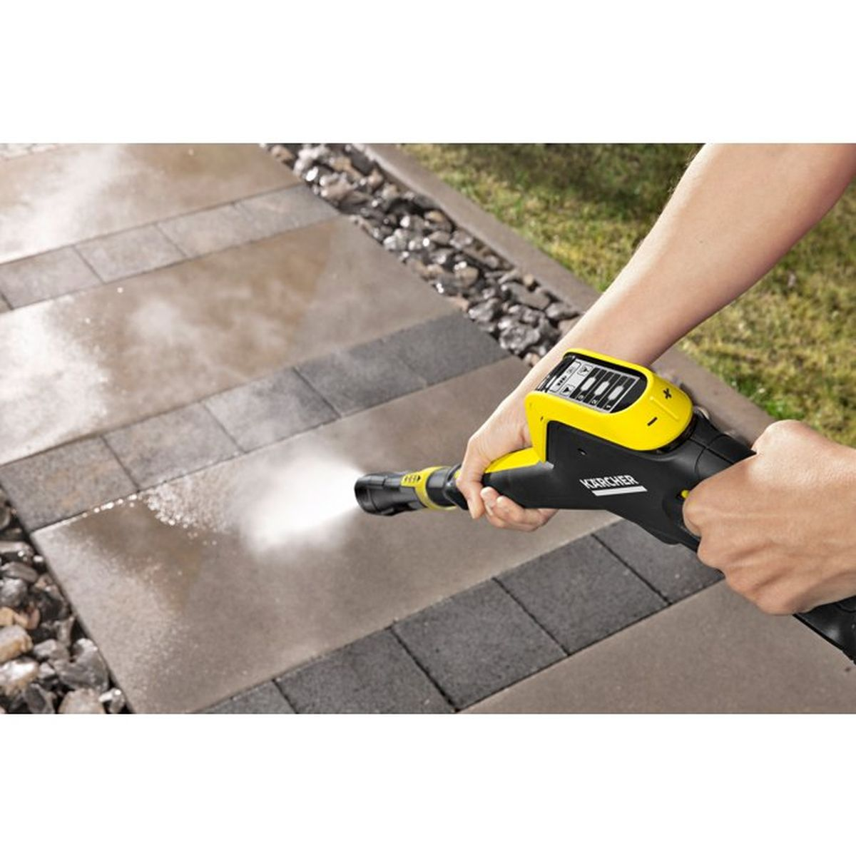 Una mano tiene un'idropulitrice Karcher gialla che spruzza acqua su un sentiero di pietra.