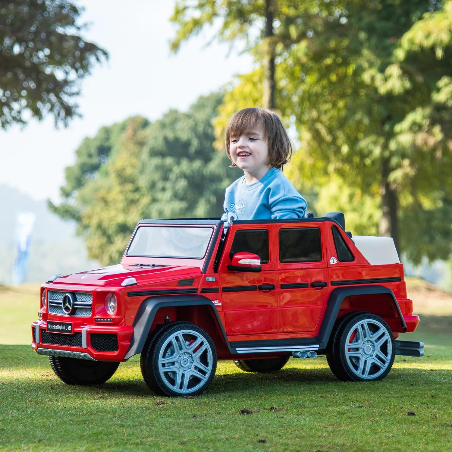 Ein lächelndes Kind sitzt in einem roten Spielzeug-Mercedes G650 auf einer Grasfläche.