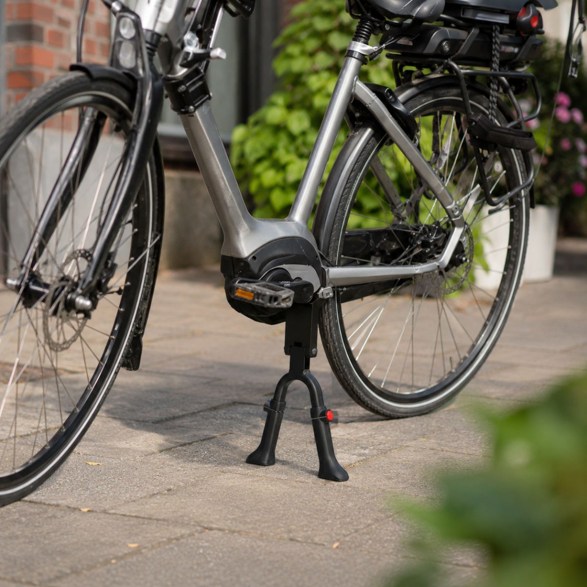 A silver e-bike stands on a stone path, with greenery in the background.