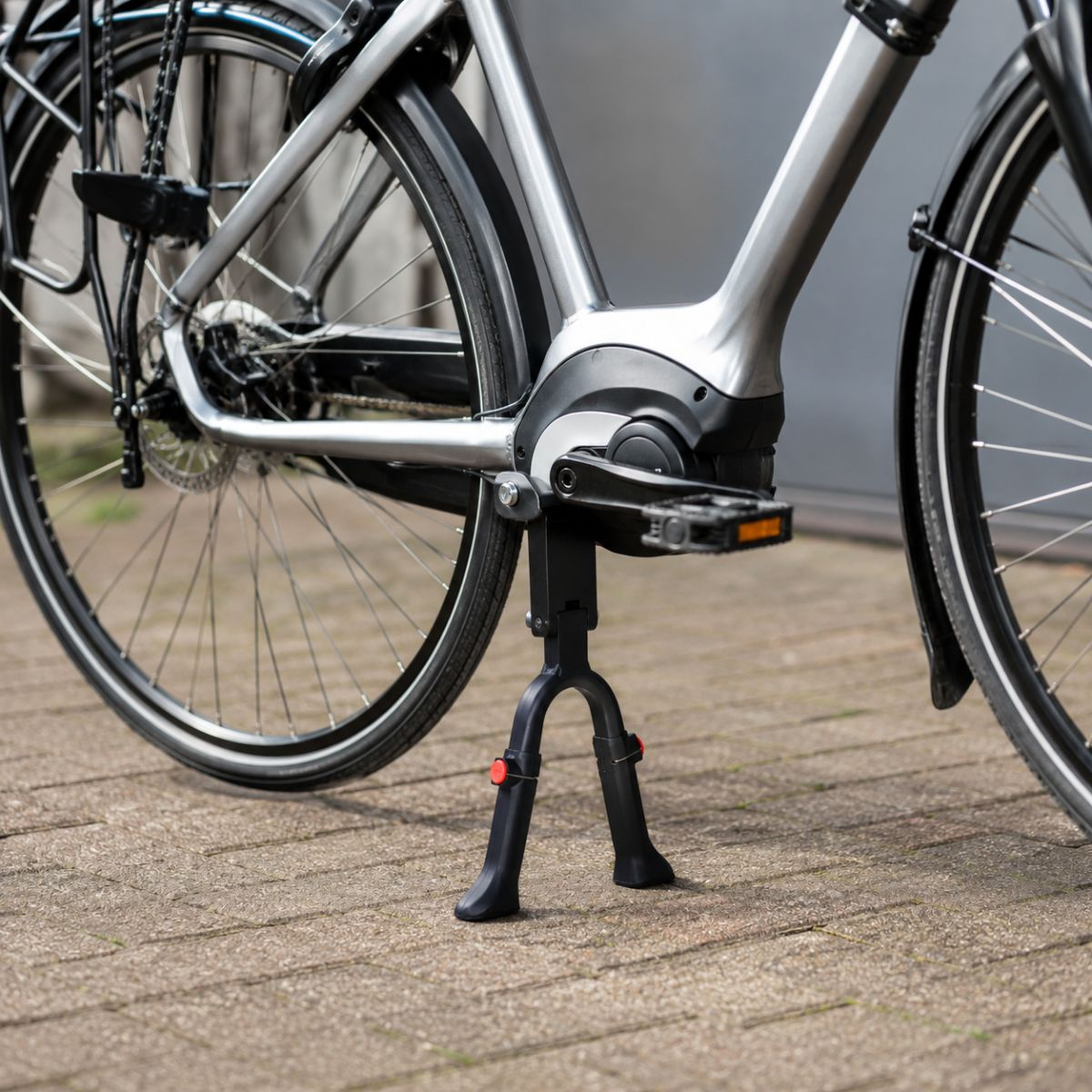 Close-up of a silver bicycle with a mid-drive motor and kickstand on cobblestone.