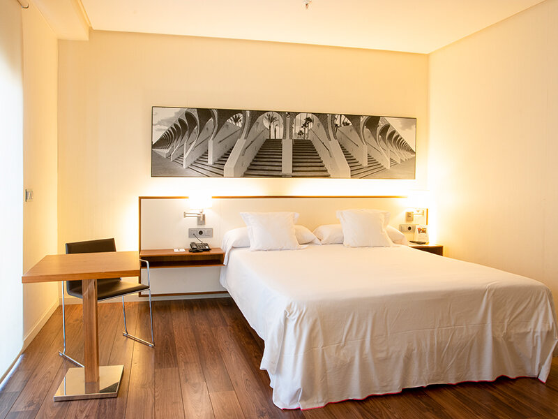 Una habitación de hotel con cama blanca, mesita de noche y escritorio.