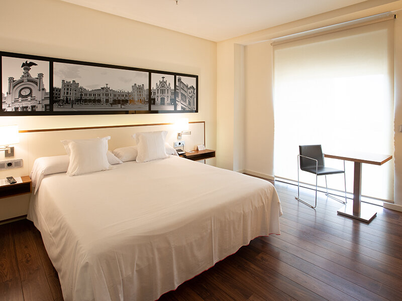 Una habitación de hotel con una cama blanca grande, silla y mesa negras junto a una ventana.