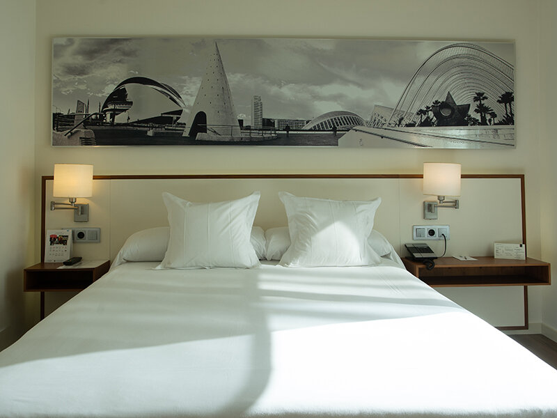 Una habitación de hotel con cama blanca, dos mesitas de noche y un mural.
